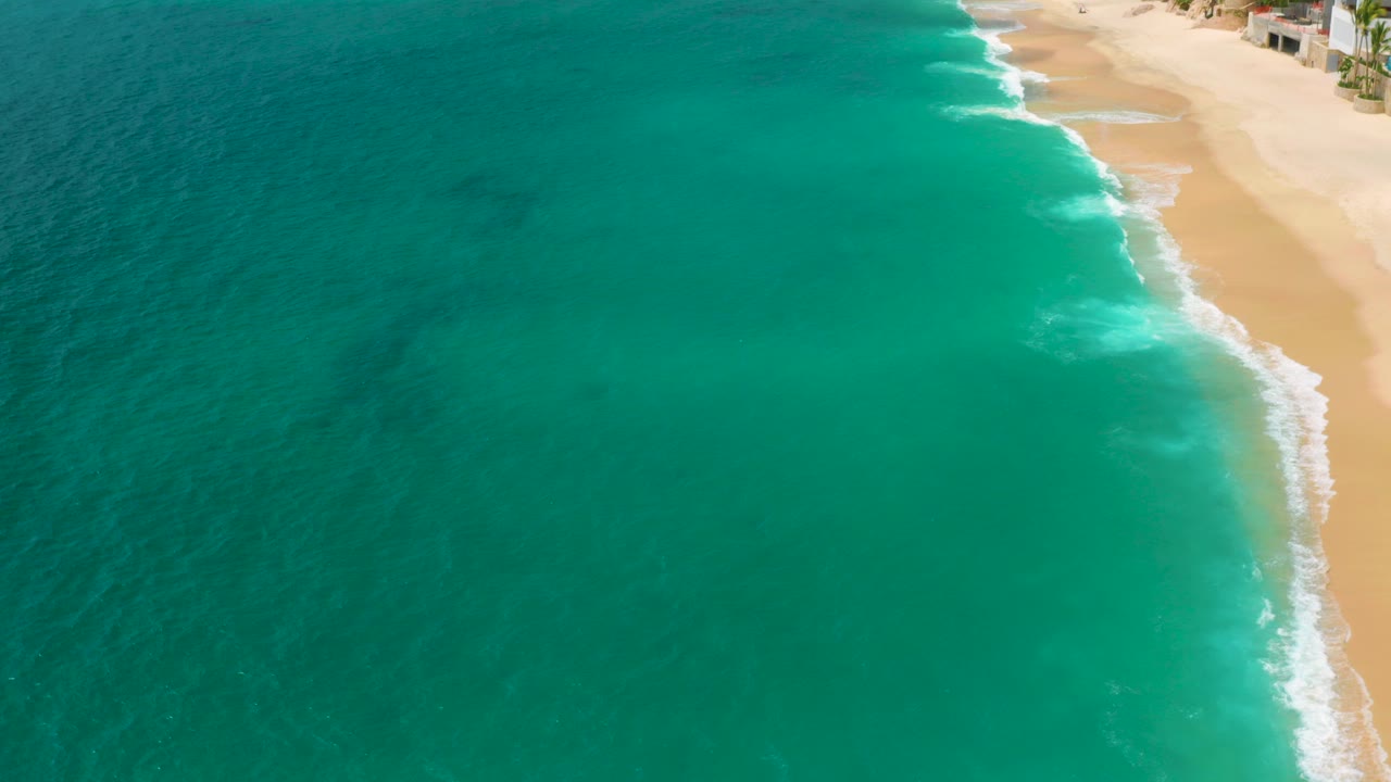 Aerial frame over Palmilla Beach Cabo, Mexico with turquoise ocean filling the left side, pale sand on the right, white foam along the shore, and no visible people.