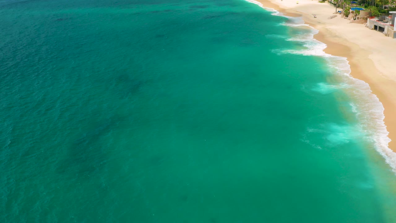 Aerial view over Palmilla Beach Cabo, Mexico with turquoise water filling most of the frame, white surf at the right edge, beachfront buildings near the top, and tiny beachgoers visible.