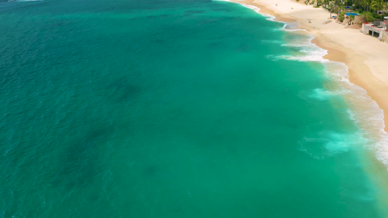Aerial frame of Palmilla Beach Cabo, Mexico showing a broad curve of turquoise water, light sand at the upper right, white shore break, palms, and low buildings near the beach.