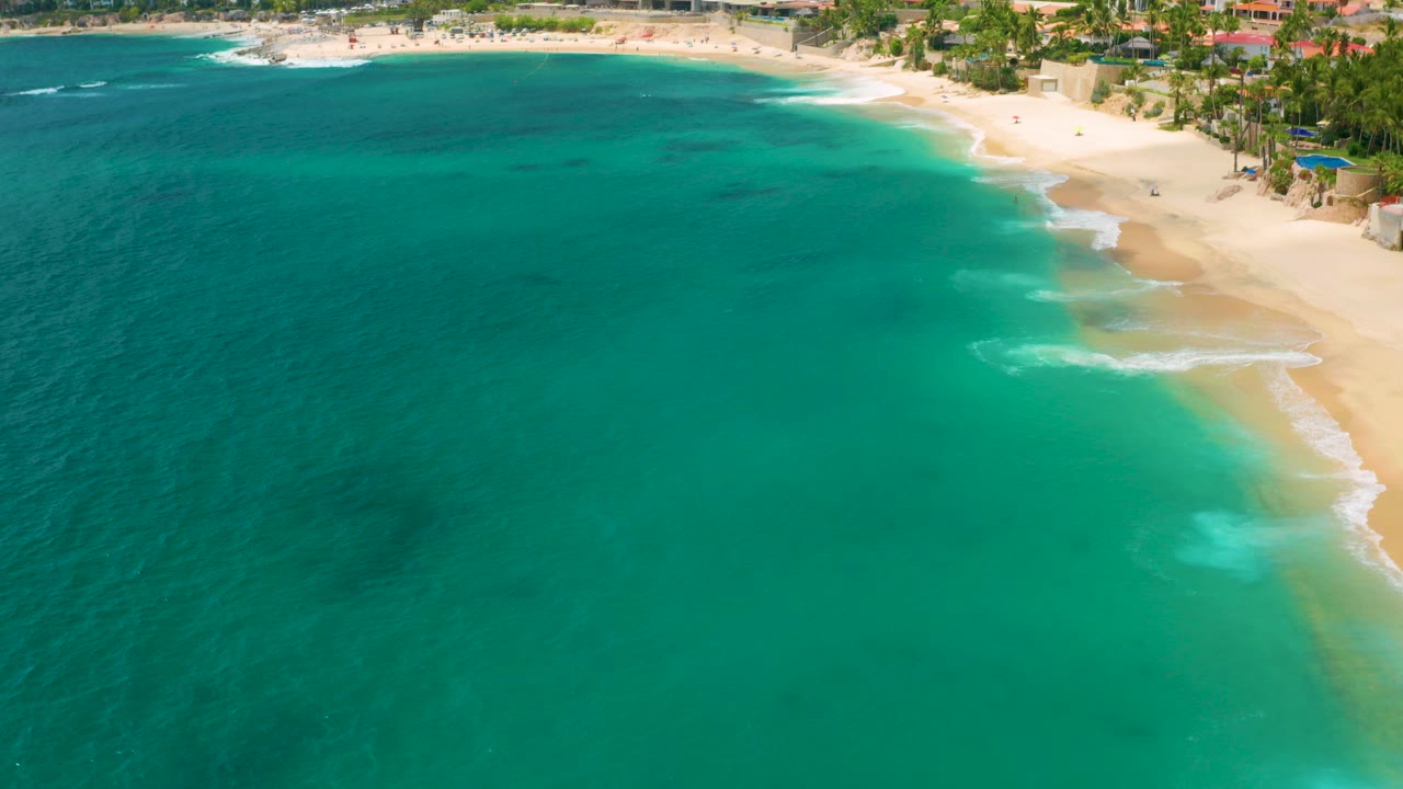 Aerial frame over Palmilla Beach Cabo, Mexico with turquoise water dominating the foreground, pale sand along the upper right, resort buildings behind palms, and no visible people.