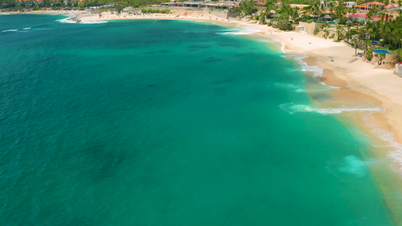 Wide aerial frame of Palmilla Beach Cabo, Mexico with the sandy beach across the top, turquoise water below, foamy surf near shore, palms, and no visible people.