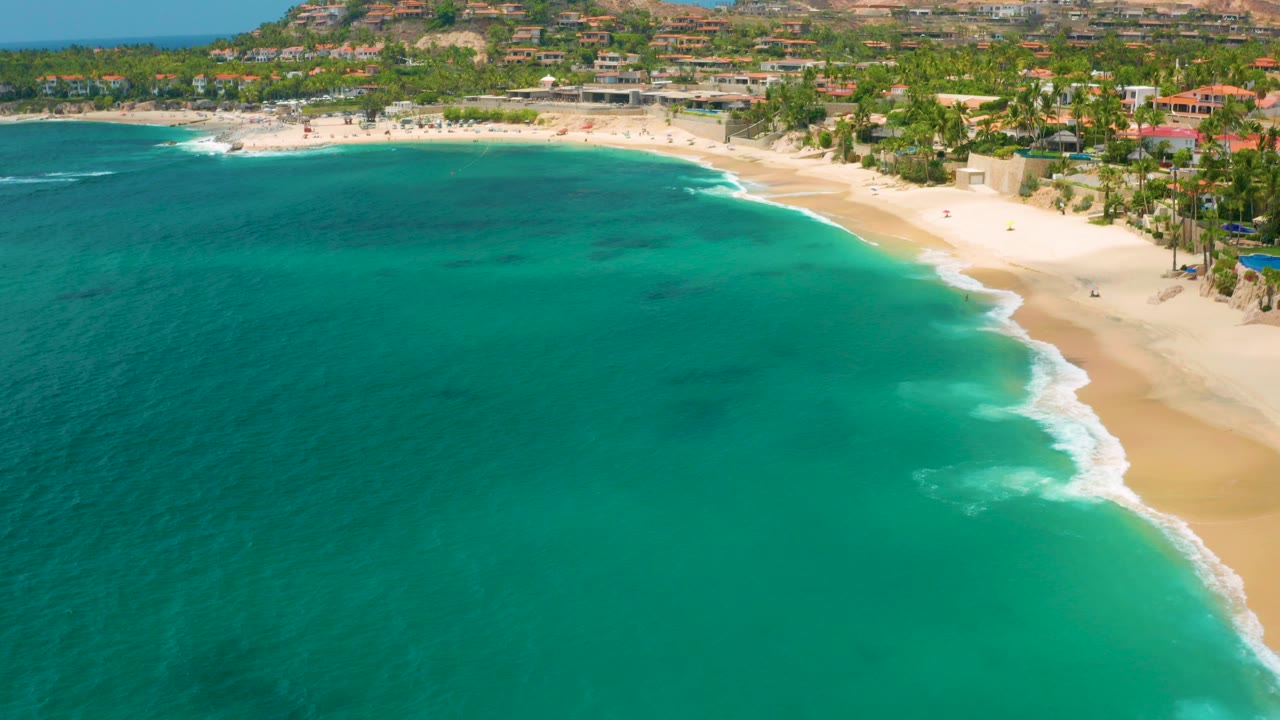 Opening aerial frame over Palmilla Beach Cabo, Mexico, with turquoise water filling the foreground, a pale sand curve at right, palm trees and hillside buildings beyond, and no visible people.