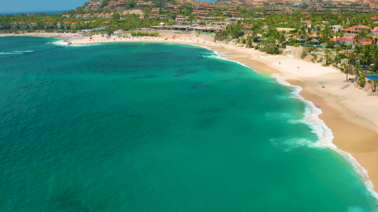 Aerial view at 0:01 over Palmilla Beach Cabo, Mexico, with more open sea across the lower frame, surf tracing the right shoreline, palms behind the sand, and no visible people.