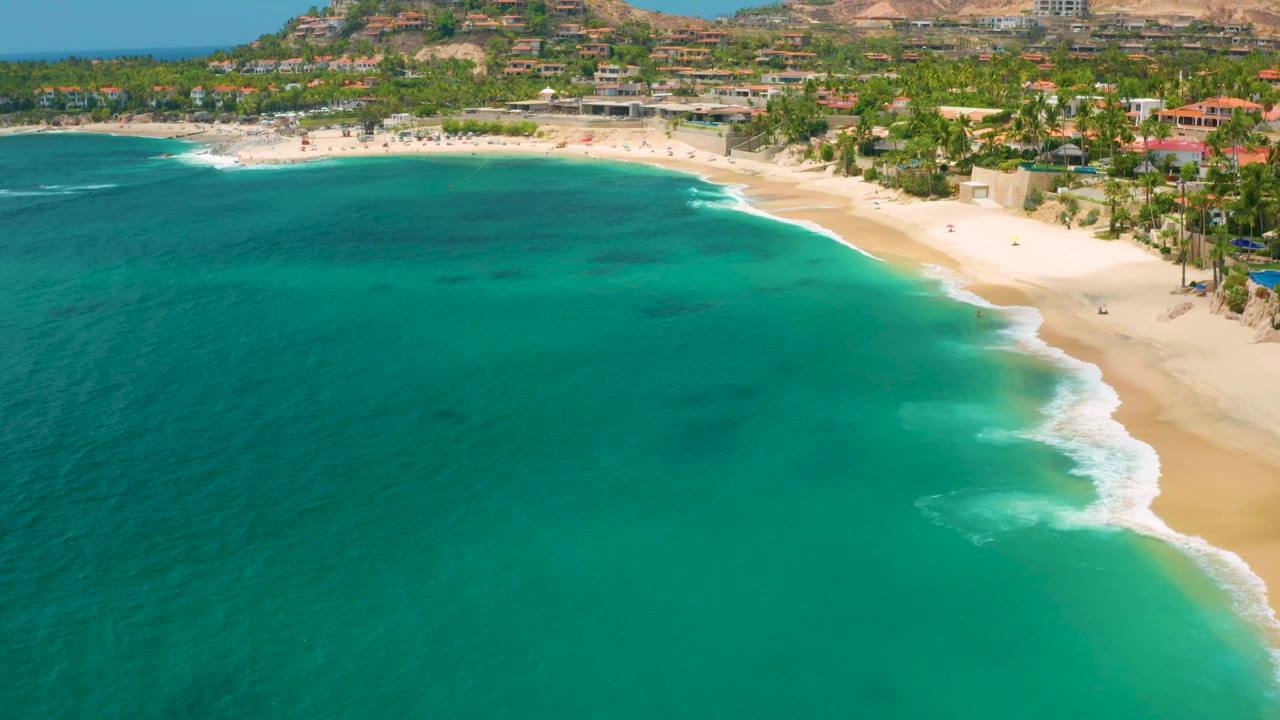 Aerial frame from Palmilla Beach Cabo, Mexico, showing blue-green water across most of the image, a foamy surf edge on the right, palm-lined sand above it, hillside buildings in back, and no visible people.