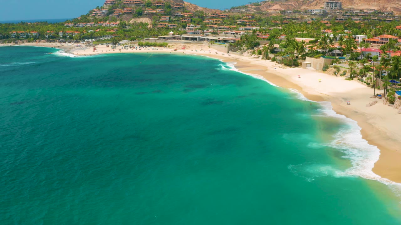 Aerial coastline view with turquoise bay, pale sand, surf, palms, and hillside homes.