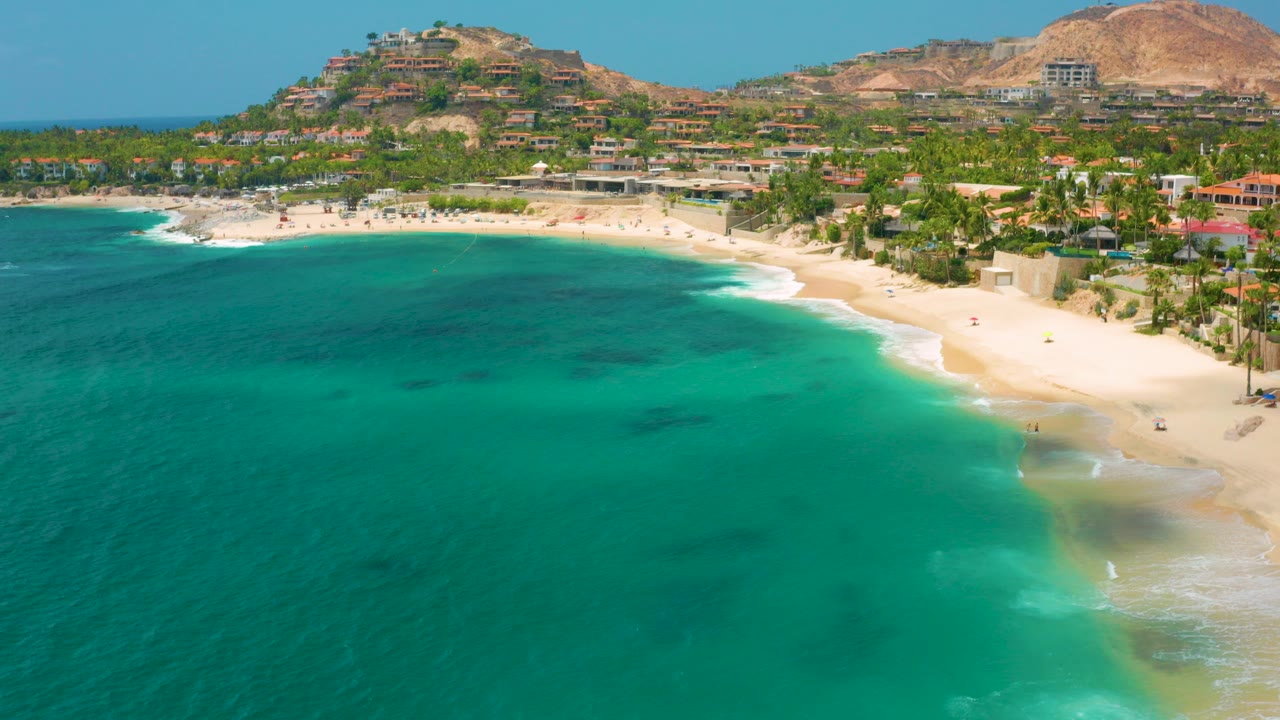A wide aerial frame over Palmilla Beach Cabo, Mexico shows turquoise water in front, a curved pale beach, scattered tiny beachgoers, palms, low buildings, and dry hills beyond.