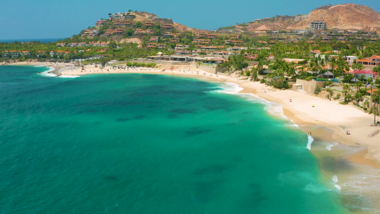 An aerial view of Palmilla Beach Cabo, Mexico shows the bay occupying most of the frame, with the beach and palms running along the right side and hillside buildings in back.