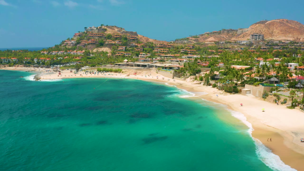 Palmilla Beach Cabo frame one second in, showing the turquoise bay spread across the lower frame, the beach running along the right edge, palms and low buildings near shore, dry hills beyond, and no discernible people.