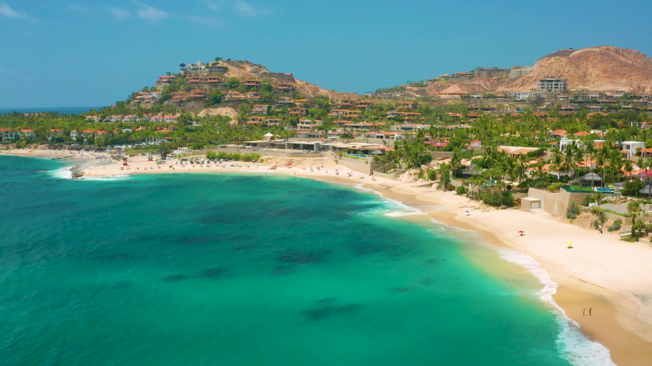 Palmilla Beach Cabo aerial frame with green-blue water across the foreground, a longer stretch of sand visible to the right, clustered palms, hillside structures, dry mountains, and no discernible people.