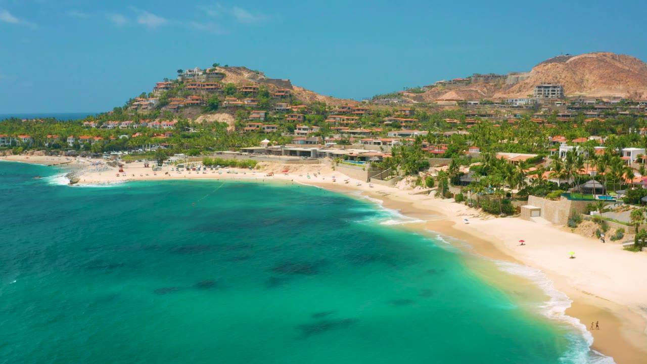 Drone view of curved beach, turquoise sea, palms, hillside residences, and mountains under blue sky.