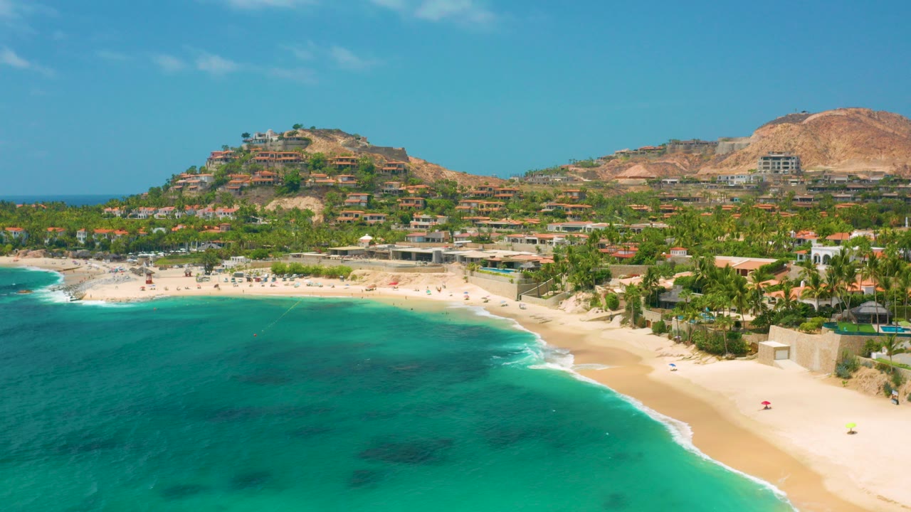 Aerial view of Palmilla Beach Cabo, Mexico with turquoise water in the foreground, a curved sand beach, palms, hillside buildings, and no visible people.