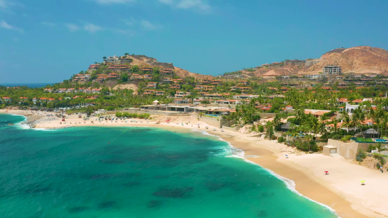Palmilla Beach Cabo aerial frame showing more open turquoise water across the foreground, pale sand along the shore, palm-lined buildings, dry hills, and no visible people.