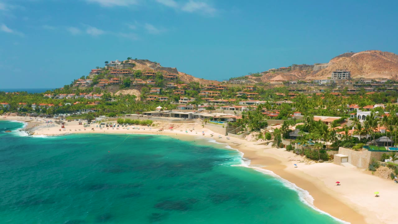 Aerial view over Palmilla Beach Cabo with the beach curve centered, turquoise water below, palms and light-colored buildings behind the sand, and no visible people.