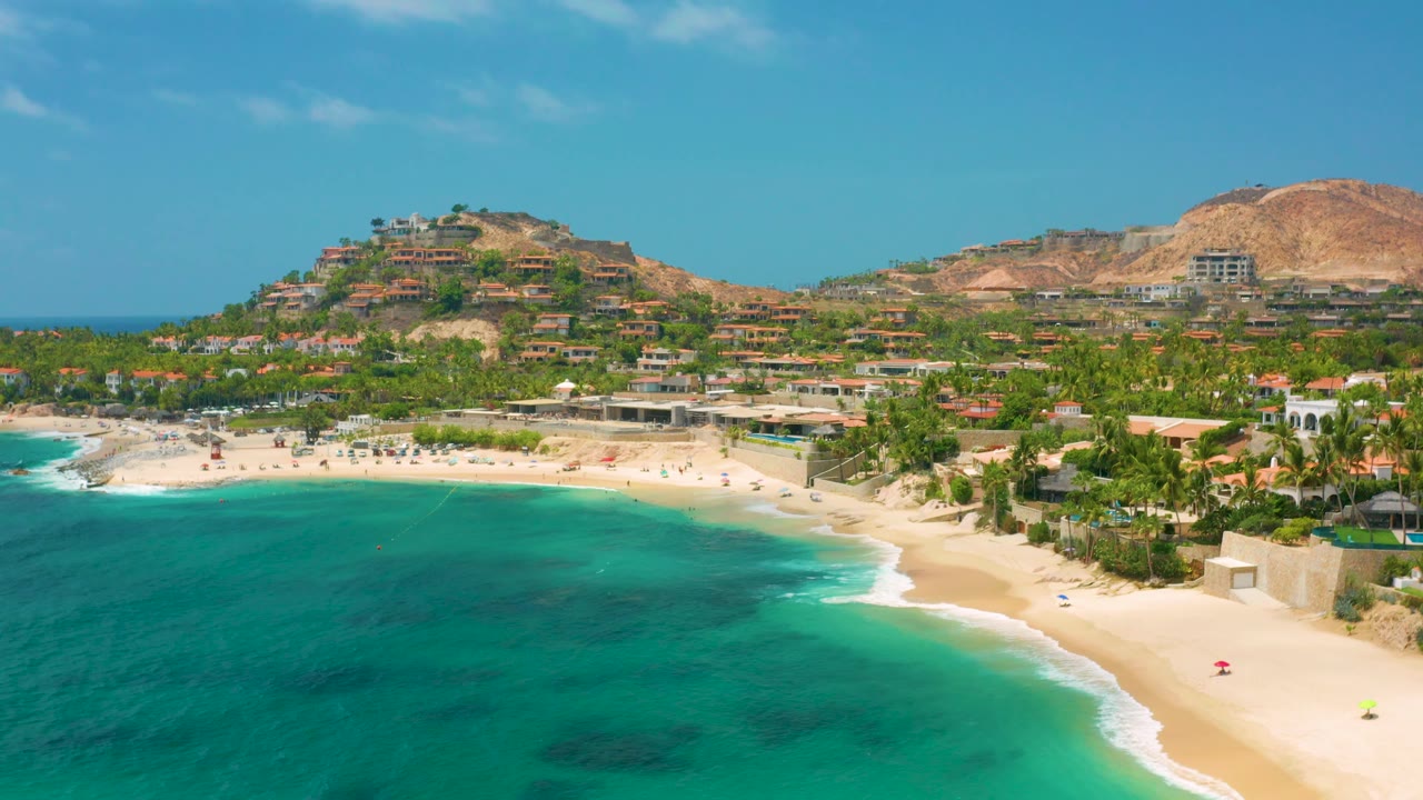 Palmilla Beach Cabo shoreline from above with a broad turquoise foreground, white surf touching the sand, dense palms, hillside structures, and no visible people.