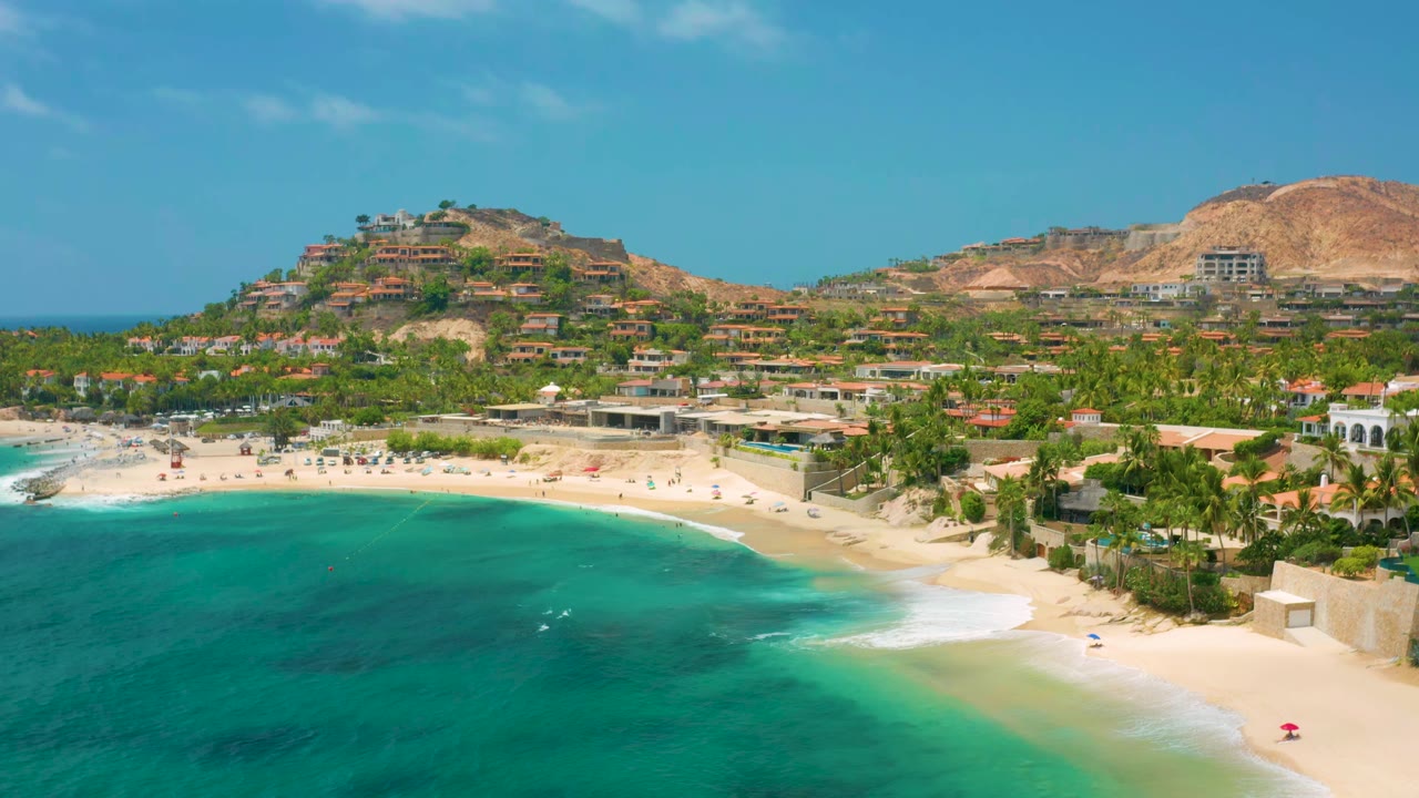 Palmilla Beach Cabo, Mexico from above with a curved surf line, beach umbrellas or loungers on the sand, dense palms, hillside buildings, and no visible people.