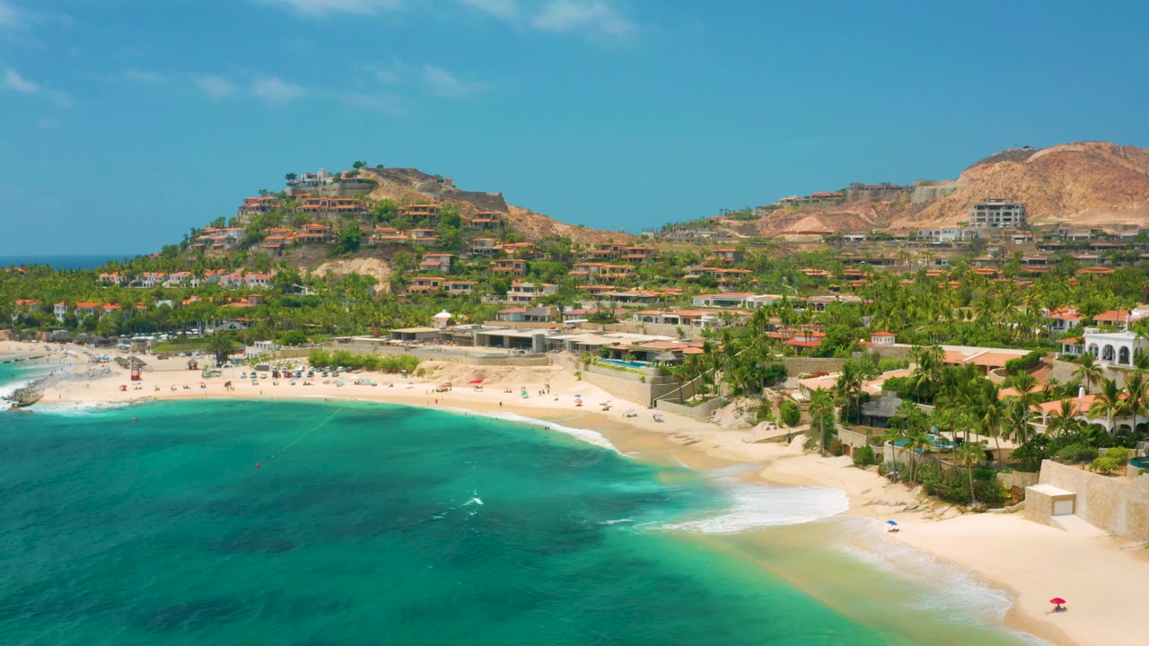 A wide Palmilla Beach Cabo, Mexico frame with clear blue-green water, a lightly occupied-looking sand area, palms, hillside structures, and no visible people.