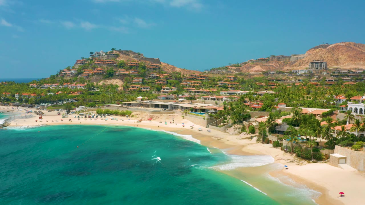 Elevated view of a sandy bay, turquoise water, palms, and hillside homes.