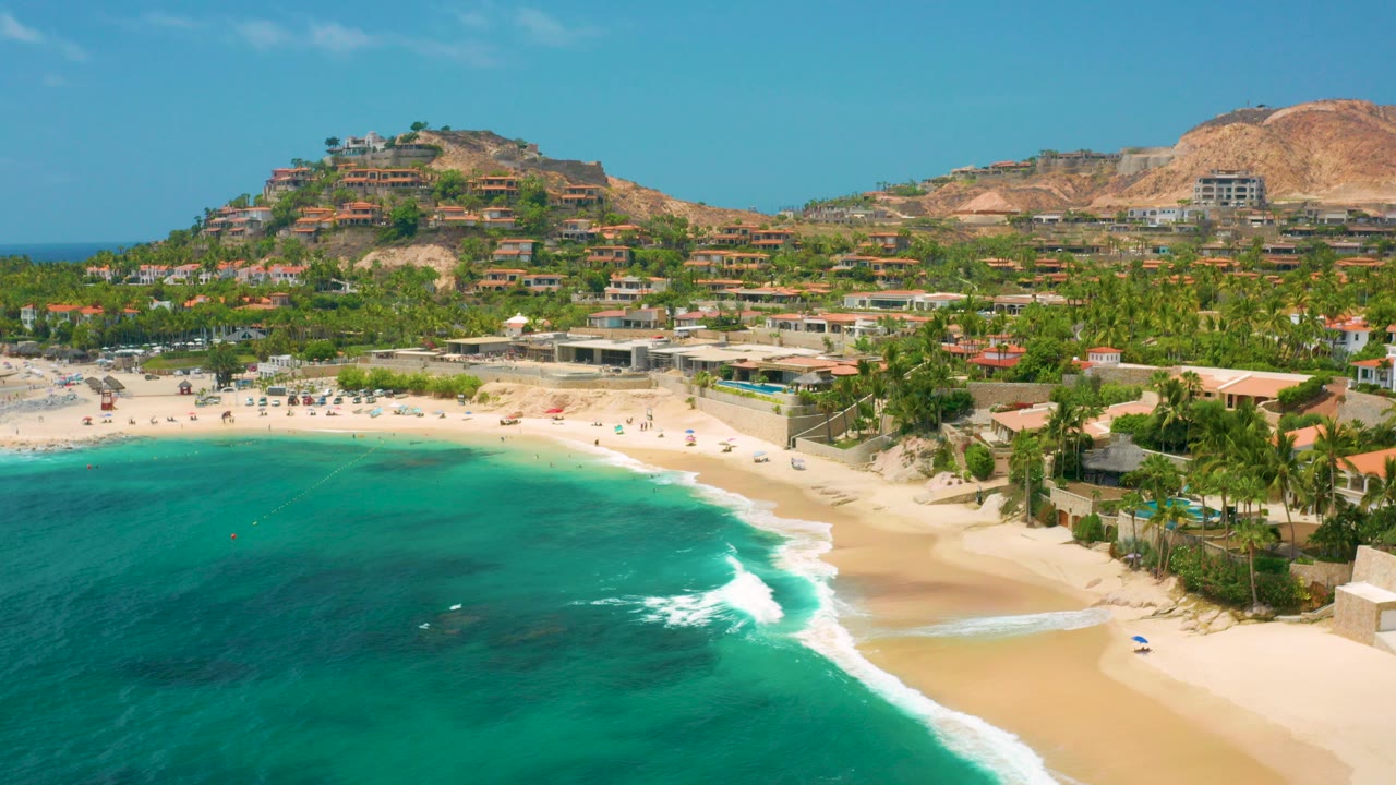 Palmilla Beach Cabo aerial frame with turquoise water in the foreground, a narrow sandy shoreline, palm trees, hillside buildings, tan slopes, blue sky, and no visible people.