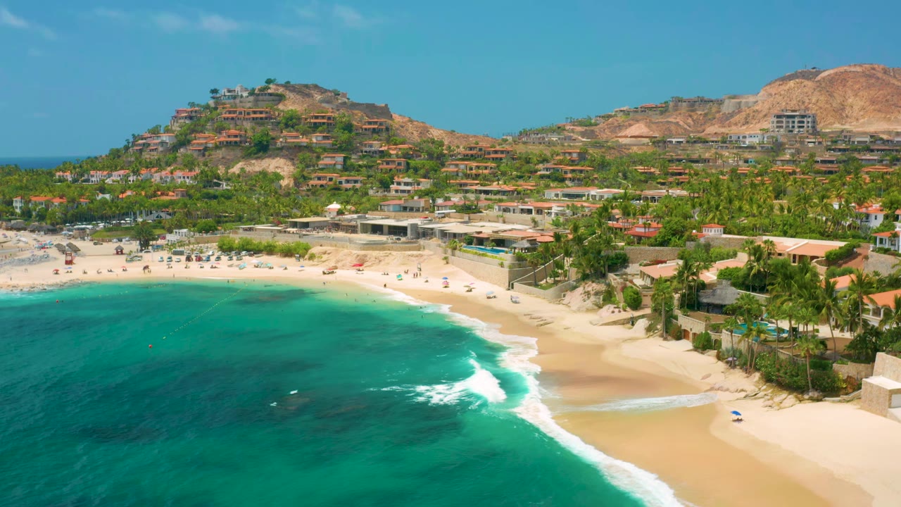 Wide aerial coastline with surf curling toward the sandy beach and hillside homes beyond.