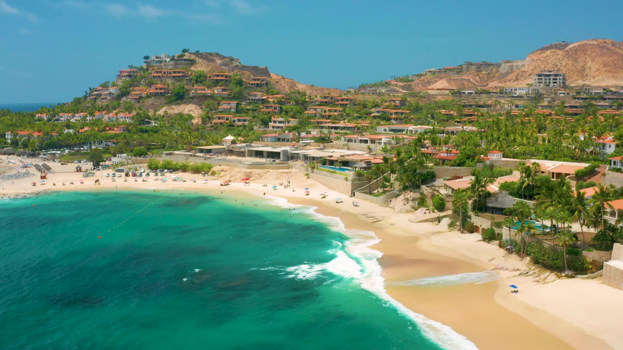 Palmilla Beach Cabo frame with the bay filling the lower half, white surf lines near the sand, palms and low buildings along the beach, terraced hillside structures, and no visible people.