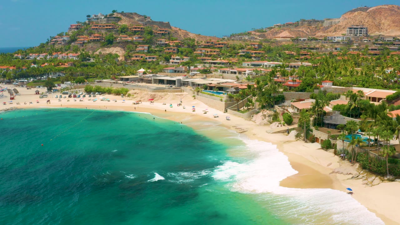 Aerial frame over Palmilla Beach Cabo, Mexico, with turquoise water in the foreground, white surf on pale sand, palms and hillside homes beyond, and no visible people.