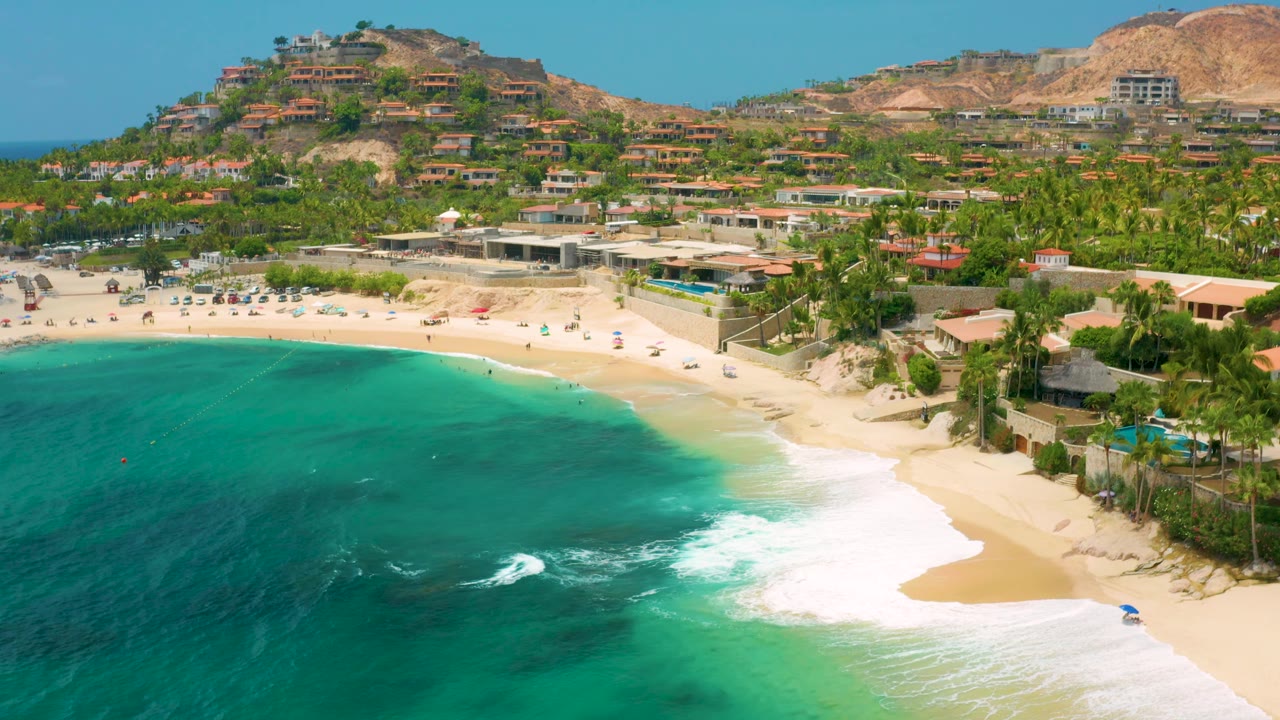 Palmilla Beach Cabo, Mexico appears from above with darker turquoise water to the left, foamy surf reaching the sand, palms and beach structures behind, and no visible people.