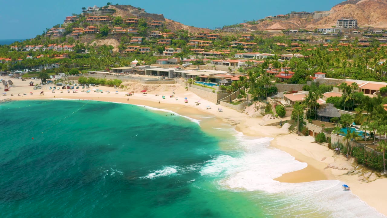 Aerial beach scene with waves washing onto sand and hillside homes in the background