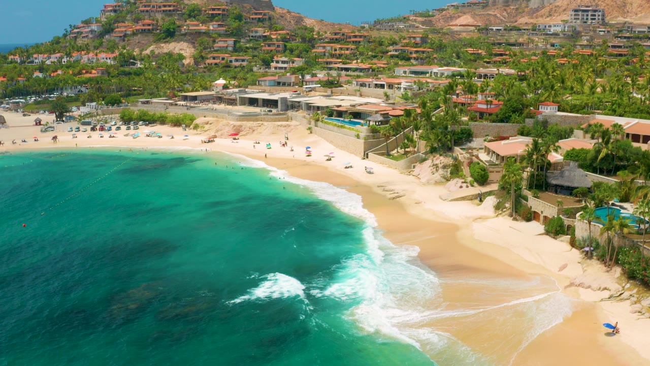 Opening aerial frame of Palmilla Beach Cabo, Mexico, with dark turquoise water in the foreground, white surf on sand, palm trees, beach umbrellas, hillside homes, and distant beachgoers.