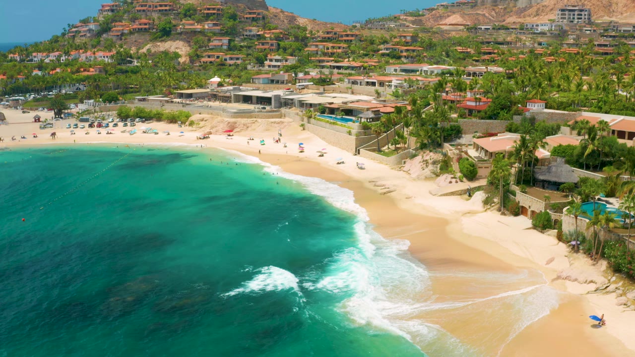 Palmilla Beach Cabo aerial view at 0:01, with the surf line curving across the lower frame, a busier strip of umbrellas and tiny beachgoers near the sand, and hillside homes behind palm trees.