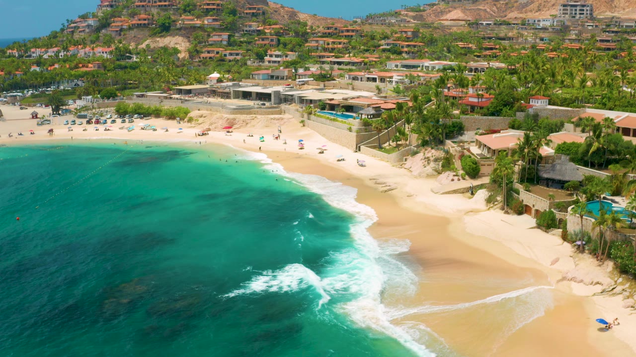 Palmilla Beach Cabo seen from above at 0:02, showing a broad turquoise bay, white water pushing up the sand, palm-lined buildings, parked cars, and distant people along the upper beach.