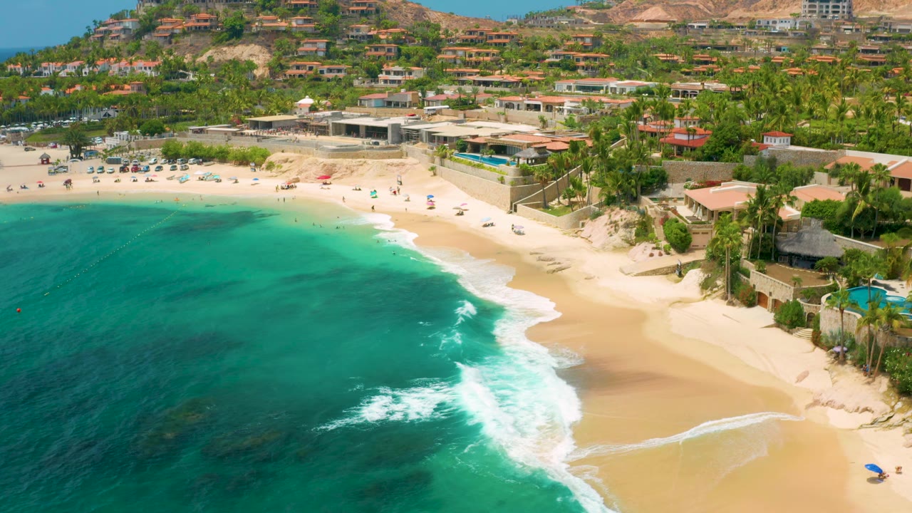 Turquoise ocean and breaking waves beside a sandy beach with palms and buildings inland.