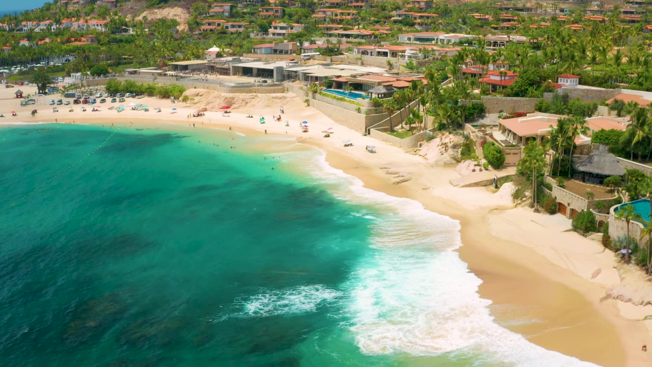 Palmilla Beach Cabo aerial still with a broad curve of turquoise water, a white wave line reaching the sand, and resort structures and hillside homes in the background, with no visible people close enough to identify.
