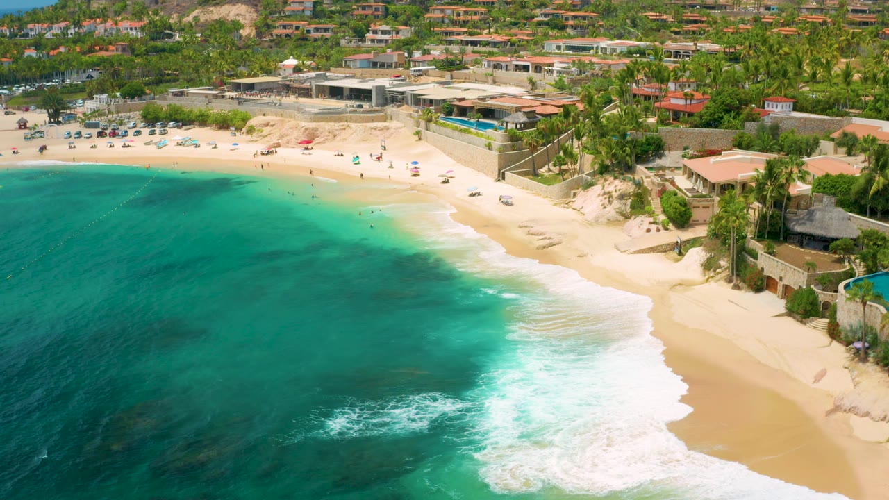 Aerial view at Palmilla Beach Cabo with darker underwater patches in the blue-green ocean, white wash along the shore, and resort and hillside buildings above the sand, with no visible people close enough to identify.