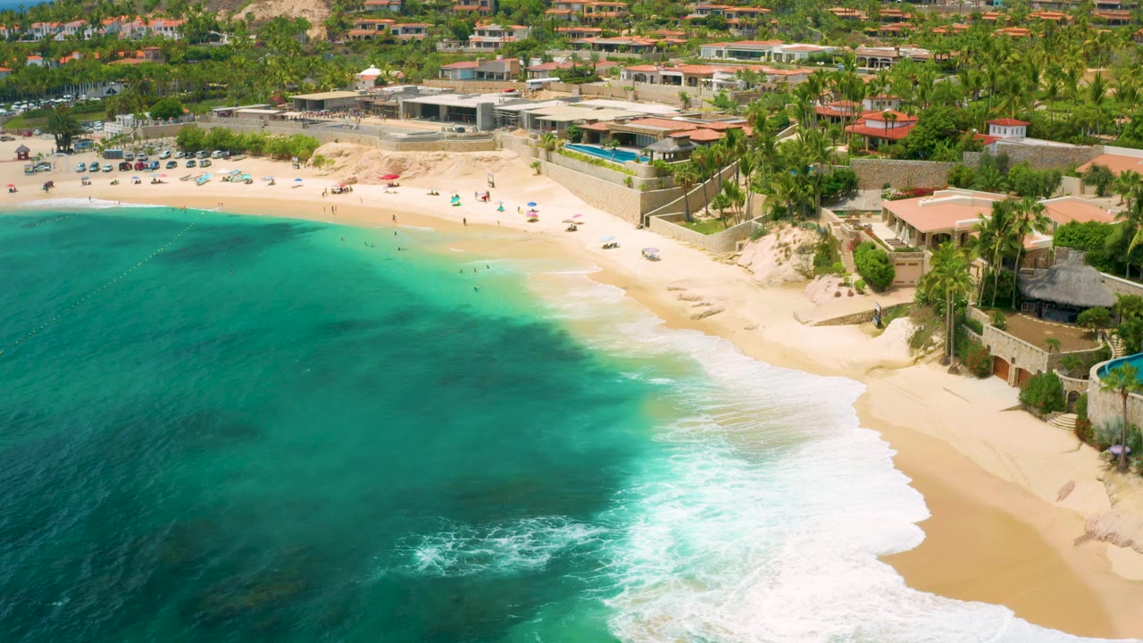 Aerial shoreline with turquoise water, white surf, sand, resort walls, and palms.