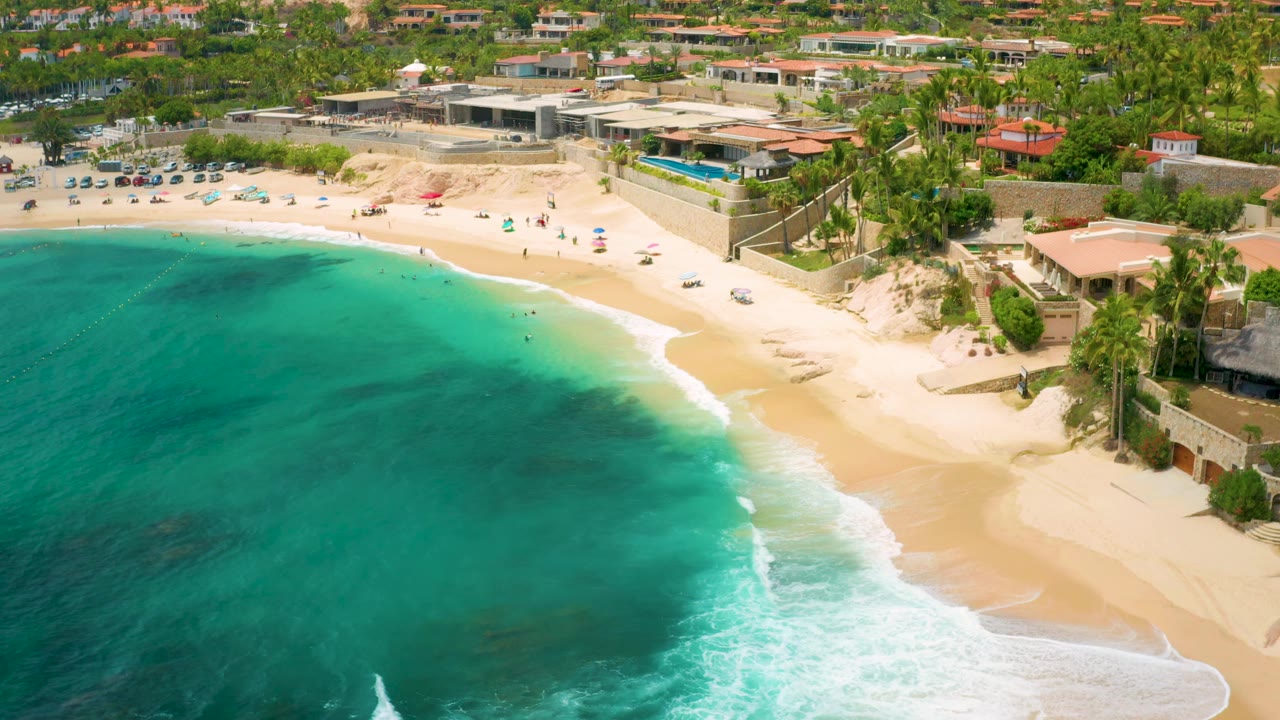 Aerial beach view with clear water, surf, umbrellas, and coastal residences.