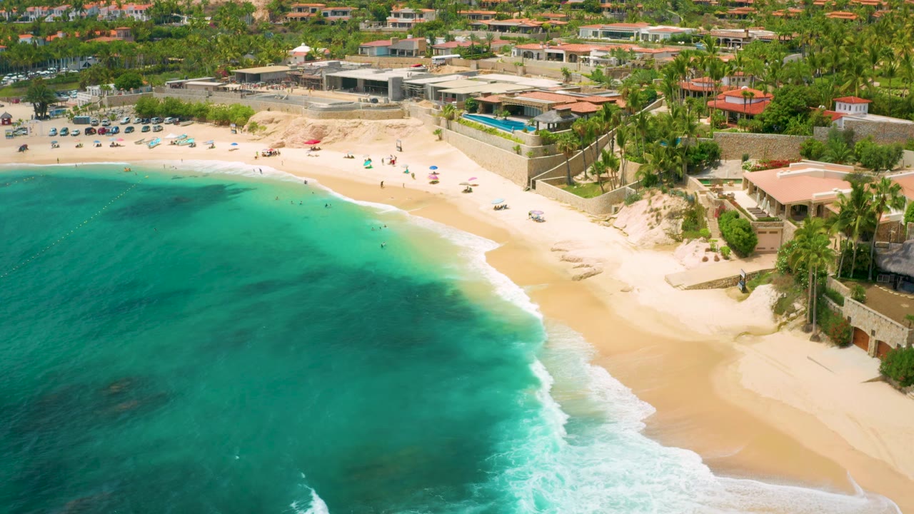 Aerial frame over Palmilla Beach Cabo, Mexico with the sea occupying most of the left side, a bright sand strip on the right, beach umbrellas near the upper shore, and palm-lined structures behind them.