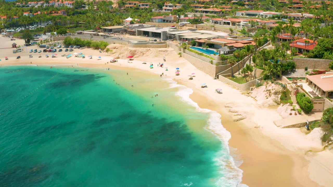 Aerial beach view with clear water, surf line, people, umbrellas, palms, and buildings.