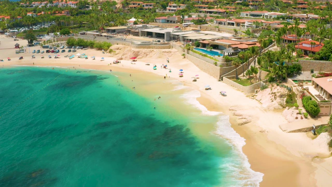Aerial view at Palmilla Beach Cabo, Mexico with deeper turquoise water across the left side, a pale sandy beach at right, tiny people near the shore, dunes, palms, and coastal buildings in the background.
