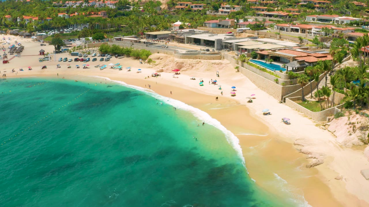 One second into the clip, the aerial view holds over Palmilla Beach Cabo with more shallow turquoise water visible, a smooth white wave line, umbrellas on the sand, and resort roofs behind the beach.