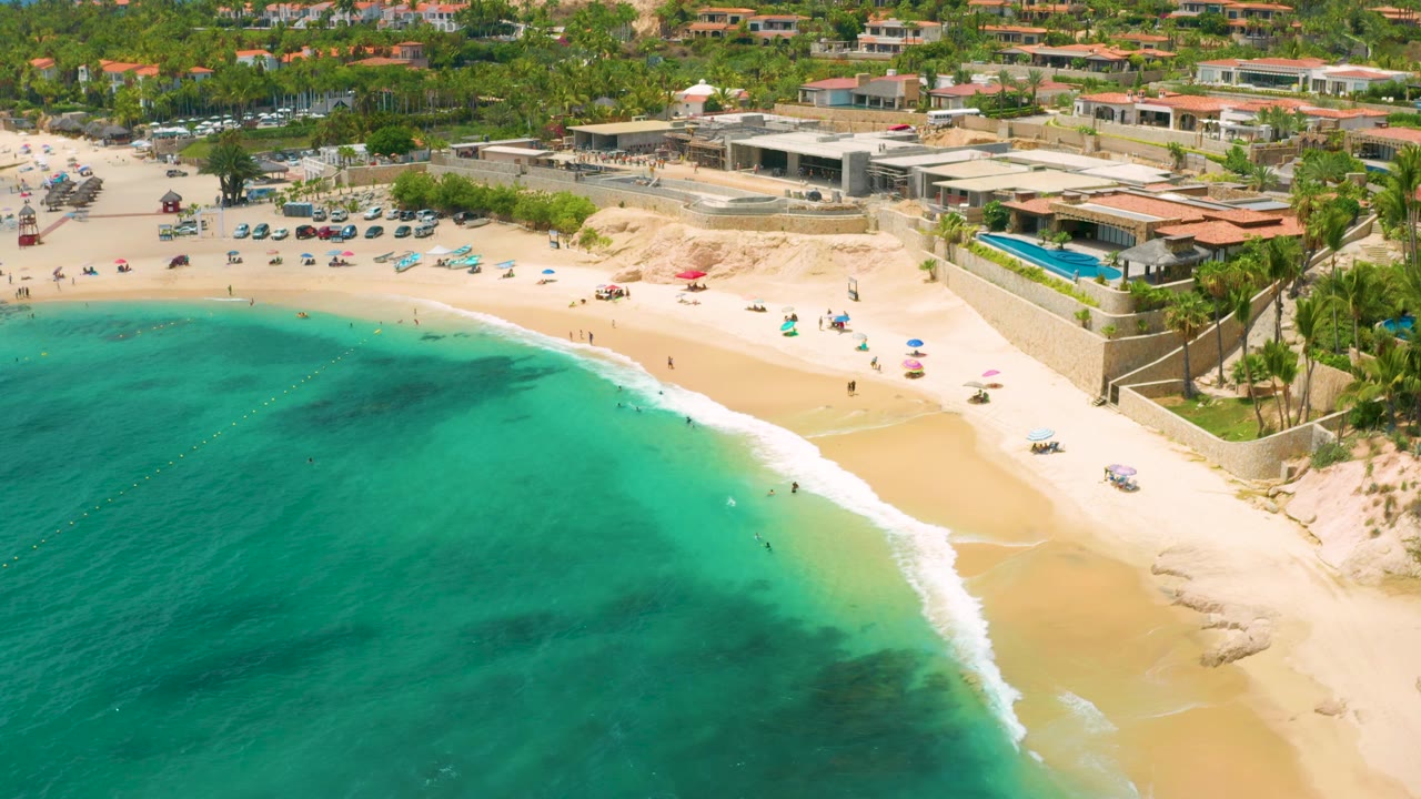 At 0:02, the frame shows Palmilla Beach Cabo from above with the shoreline curving across the image, turquoise water filling the lower left, a resort pool at right, and palm-lined buildings in back.