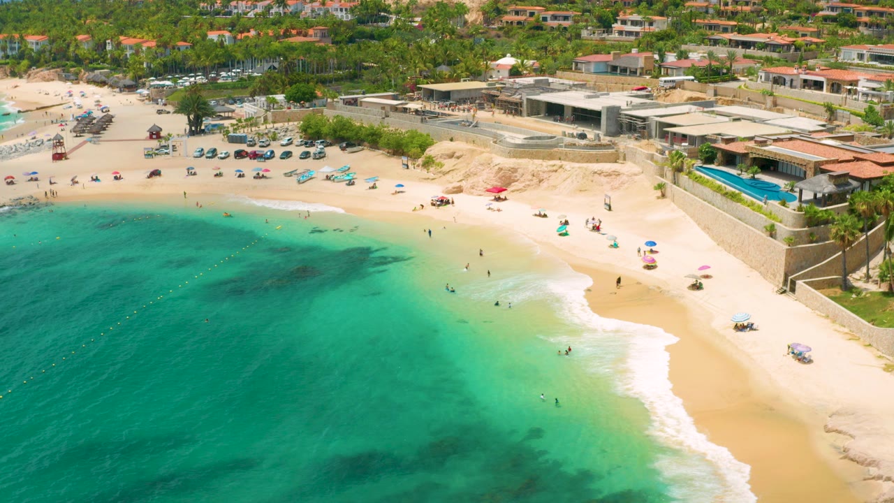 Opening aerial frame over Palmilla Beach Cabo, Mexico, with turquoise water in the foreground, pale sand, scattered umbrellas, beachgoers, parked cars, palms, and low resort buildings beyond the shore.