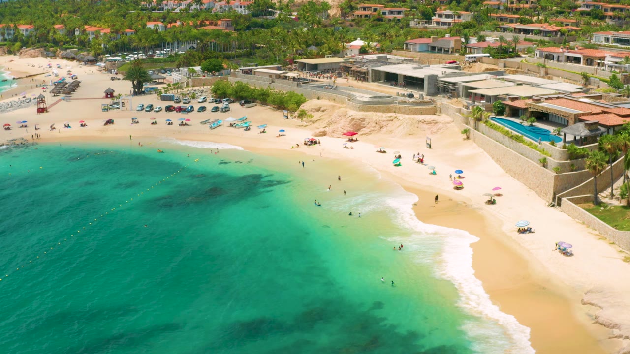 Drone view of clear water, surf, umbrellas, and beachfront buildings.