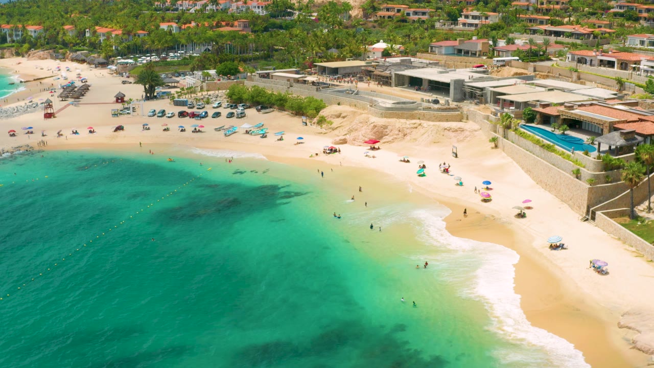 Palmilla Beach Cabo frame with more shoreline centered, showing swimmers spread through shallow turquoise water, umbrellas on the dry sand, parked vehicles near palms, and low resort buildings above the beach.