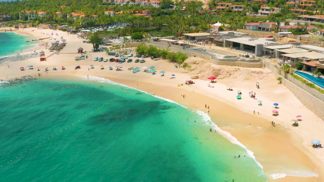 Elevated beach view with clear water, surf line, umbrellas, and palm-lined buildings.
