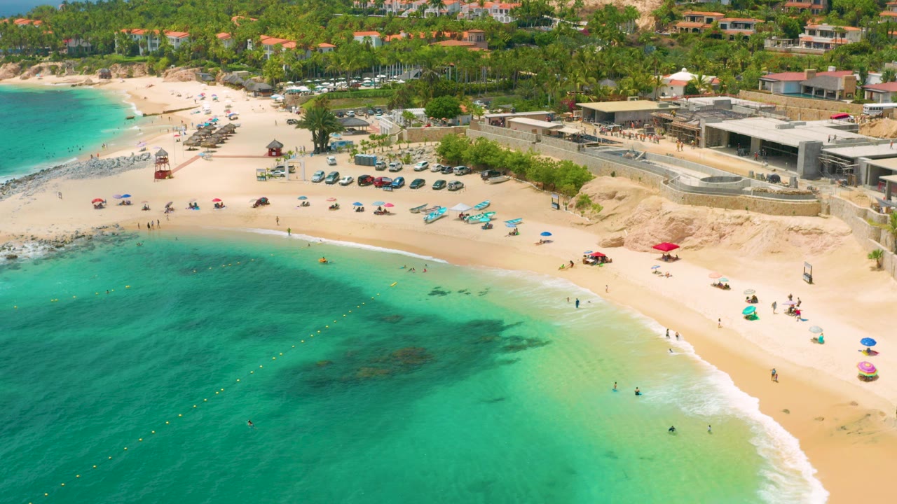 Wide aerial view at Palmilla Beach Cabo, Mexico, with turquoise water in the foreground, a curved sandy shore, scattered beachgoers, umbrellas, parked cars, palms, and low shoreline buildings.