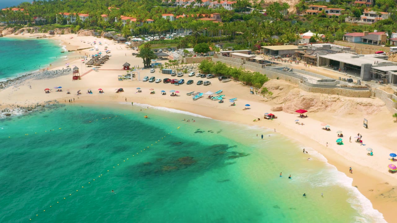 Aerial view of Palmilla Beach Cabo with more open water across the foreground, pale sand along the right edge, small people on the beach and in the surf, and palms and buildings beyond.