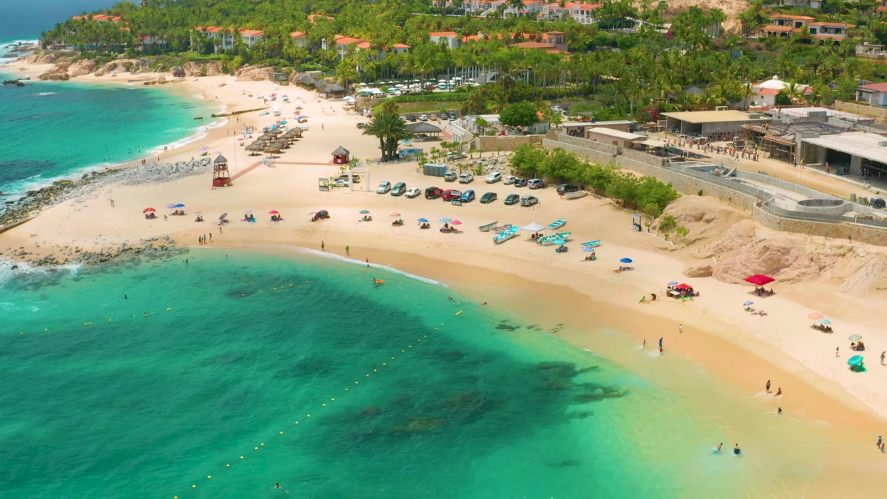 High aerial beach view with curved shoreline, umbrellas, swimmers, cars, palms, and buildings.