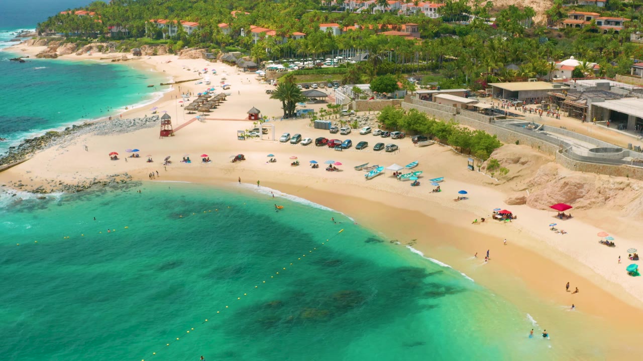 Palmilla Beach Cabo, Mexico seen from above with turquoise water across the foreground, darker reef-like patches below the surface, and a populated beach backed by palms and buildings.