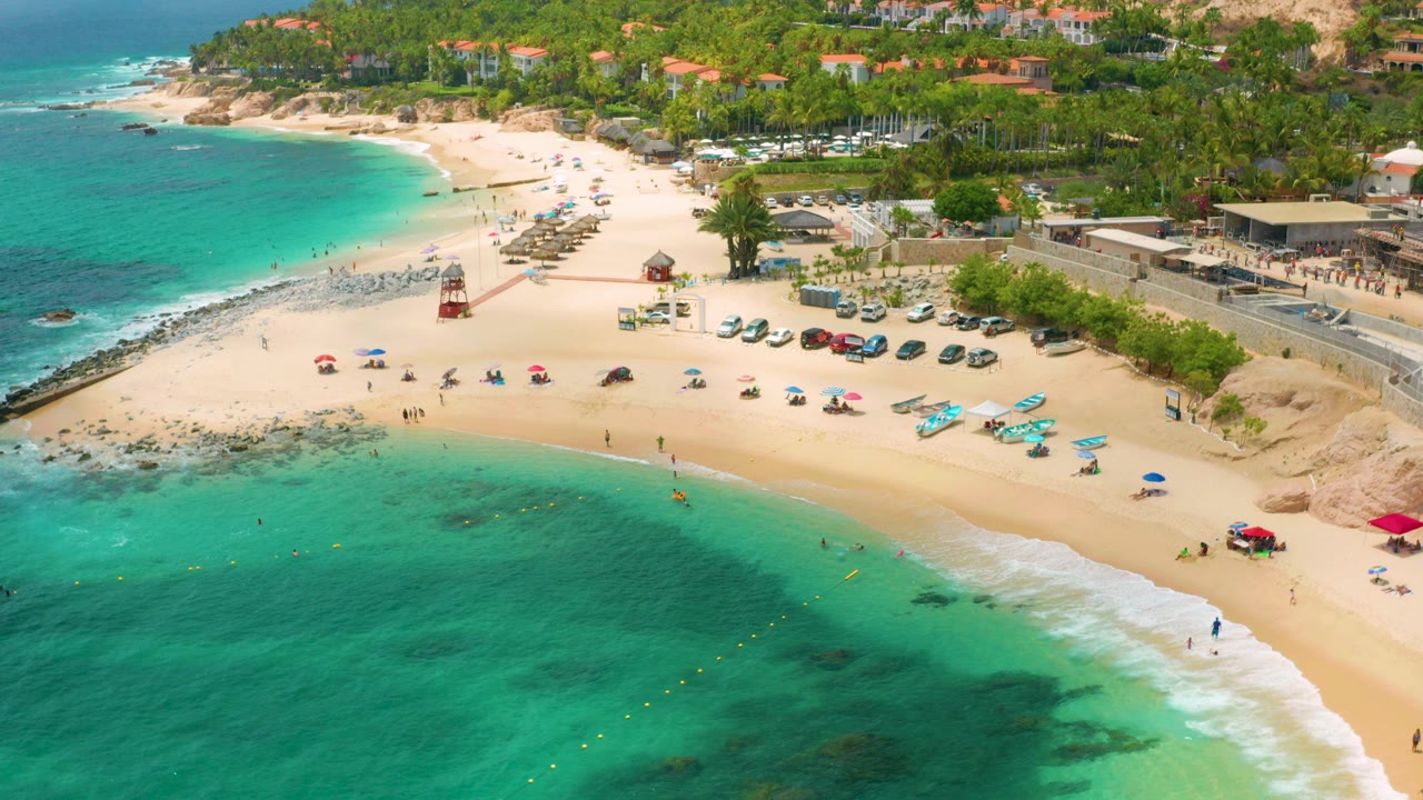 Aerial frame over Palmilla Beach Cabo, Mexico with turquoise shallows in the foreground, a red lifeguard tower near the sand, parked cars, palms, hillside buildings, and tiny visible beachgoers.
