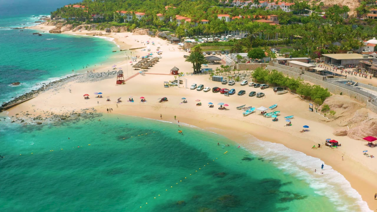 Palmilla Beach Cabo aerial still showing clear green-blue water, the curved sand edge, scattered umbrellas, a red lifeguard tower, rows of cars, palms, and small visible beachgoers.